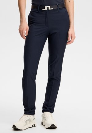 Slim-fit marineblå bukser med forreste folder, sort bælte med sølvspænde, båret med hvide sportssko.