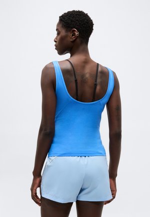 Γυναίκα με κοντά σγουρά μαλλιά που φοράει μπλε αμάνικη μπλούζα και ανοιχτού χρώματος σορτς, εμφανίζεται από πίσω με μονόχρωμο φόντο.