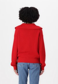 Red knitted sweater with a wide collar and ribbed cuffs, paired with light blue denim jeans. The texture appears soft and cozy.