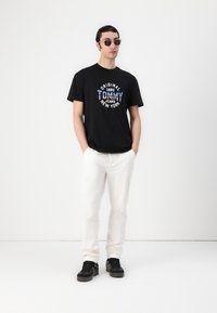 Sort t-shirt med farverig "Tommy Jeans New York" logo, båret med hvide bukser og sorte sneakers. Modellen poserer foran en ensfarvet baggrund.