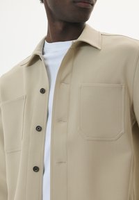 Camicia button-up beige con una texture liscia, una tasca singola e bottoni neri, sovrapposta a una maglietta bianca a girocollo.