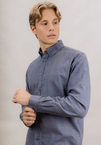 Blå rutete langermet skjorte med knappkrage, med blå knapper og myk tekstur. Modell justerer mansjett.