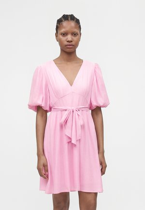 Žena s vrkočmi, ktorá má na sebe svetloružové šaty s výstrihom do V, nadýchanými rukávmi a stiahnutým mašľou v páse, stojí pred jednofarebným pozadím.