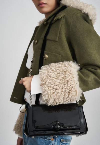 Saco de mão de pele sintética preta com uma alça de corrente, formato retangular e um detalhe de fecho. O modelo usa um casaco de lã verde com um punho de pele.