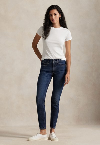 Donna con lunghi capelli ricci che indossa una maglietta bianca, jeans skinny scuri, sneakers bianche e orecchini a cerchio d'oro su uno sfondo neutro.