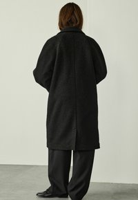 Person with shoulder-length brown hair wearing a long black textured coat, black wide-leg pants, and black shoes, standing indoors.