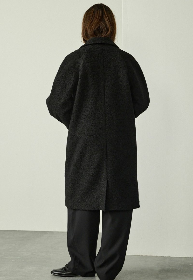 Person with shoulder-length brown hair wearing a long black textured coat, black wide-leg pants, and black shoes, standing indoors.