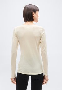 Γυναίκα με κοντά σκούρα μαλλιά που φοράει εφαρμοστό μακρυμάνικο ανοιχτό μπεζ τοπ, φαίνεται από πίσω πάνω σε απλό λευκό φόντο.