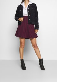 Vrouw in een zwarte corduroy jas, witte overhemd met kraag, rode en marineblauwe gestreepte A-lijn rok en zwarte enkellaarzen, staand op een grijze vloer.