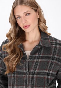 Grå rutig flanellskjorta med röda och svarta detaljer, med krage och knäppning, mjuk textur. Modellen bär en beige topp under.