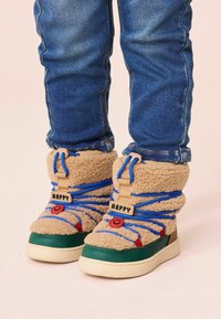 Bottes en fourrure marron avec accents bleus, détail de smiley rouge et semelles en caoutchouc vert. Elles présentent une sangle de marque et un système de laçage durable.