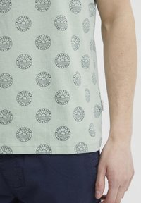 Lys grønn bomulls T-skjorte med et mønster av sirkulære svarte grafikker. Korte ermer, rund hals, med et lite logo-merke på siden.