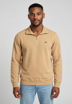 Homme portant un pull beige à fermeture éclair quart, avec un petit logo crocodile vert sur la poitrine et un jean bleu clair, debout devant un fond clair.