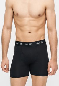 Ανδρικός κορμός με μαύρα εσώρουχα Hollister, δείχνει καθορισμένους κοιλιακούς και χέρια σε απλό ανοιχτό φόντο.