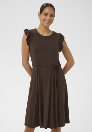 Femme souriante, portant une robe marron sans manches avec des épaules à volants et une ceinture à la taille, debout devant un fond blanc uni.