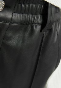 Schwarze Lederhose mit einer glatten Textur, gerafftem Bund, Seitentaschen und einem metallischen Knopfakzent nahe dem Bund.