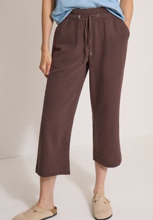 Person iført løse, brune, cropped bukser med snøreliv, lyseblå skjorte og beige slip-on sko står i et neutralt indendørs rum.