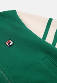 Felpa verde con accenti bianchi, dotata di una chiusura a zip frontale, texture morbida e un piccolo logo multicolore sul lato sinistro.