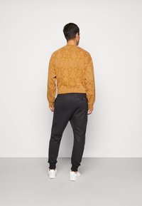 Brun mønstertrøje med rund hals, parret med sorte joggers og hvide sneakers, set bagfra.