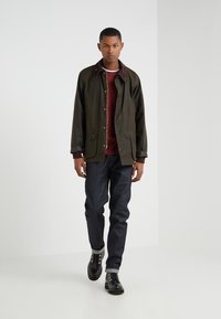 Olive green jacket with brown collar and buttons, paired with a maroon sweater and dark denim jeans. Black lace-up boots with silver eyelets.