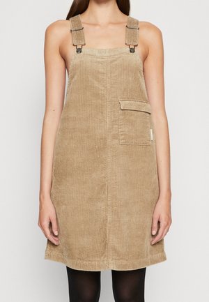 Vrouw draagt een beige corduroy tuinbroekjurk met brede bandjes en een voorzak, gecombineerd met zwarte panty's.