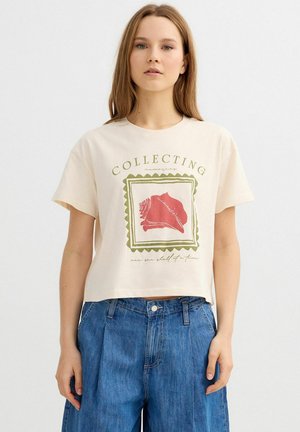 Joven mujer con una camiseta color crema con un gráfico de una concha roja y el texto «COLLECTING memories», combinada con vaqueros azules de tiro alto.