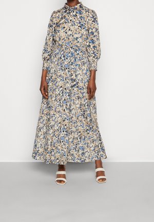 Femme portant une longue robe fleurie avec des motifs bleu, beige et noir, boutonnée devant, taille cintrée et sandales blanches à brides tressées.