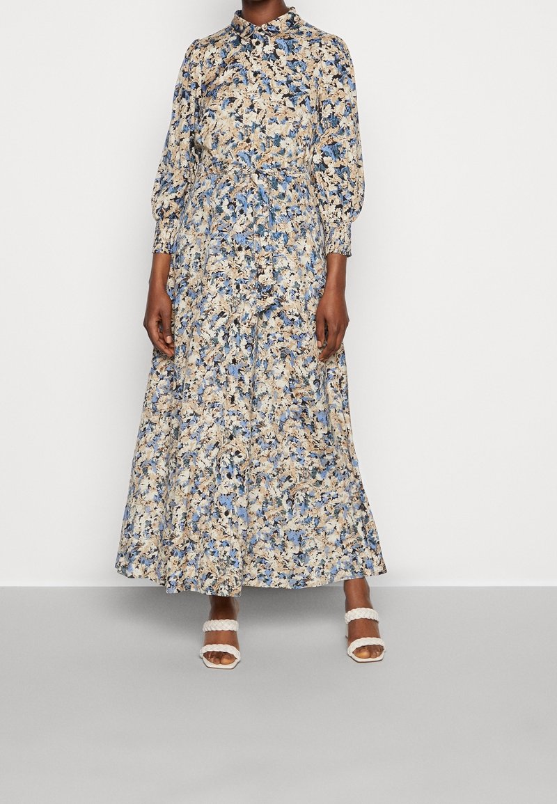Femme portant une longue robe fleurie avec des motifs bleu, beige et noir, boutonnée devant, taille cintrée et sandales blanches à brides tressées.