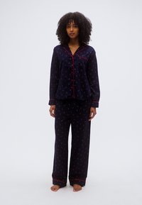 Ensemble de pyjama bleu marine avec passepoil rouge, comprenant un haut boutonné et un pantalon ample, orné de petits motifs d'ancres colorées. Tissu en mélange de coton.