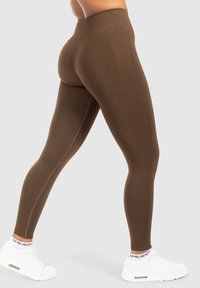 Leggings marroni a vita alta realizzati in tessuto elastico, con una texture liscia e cuciture laterali, abbinati a scarpe da ginnastica bianche.