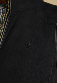 Melna vilnas jaka ar rāvējslēdzēju, kas izceļas ar diskretu logo uz krūtīm un kontrastējošiem ādas akcentiem gar rāvējslēdzēju.