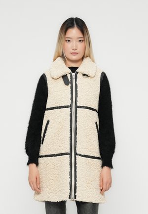 Jeune femme de face, portant un gilet sans manches en sherpa crème avec des bordures noires sur un haut à manches longues noir, devant un fond uni.