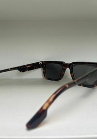 Tortoiseshell zonnebril met vierkante glazen. Donkergrijze getinte glazen, solide zwarte armen met een glanzende afwerking en metalen accenten aan de zijkant.