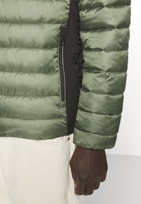 Gros plan d'une personne portant une veste matelassée verte avec une fermeture éclair noire sur le côté et un pantalon blanc, montrant le bras et la main droite.