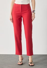 Pantalons taillés rouges à coupe droite, avec des plis à l'avant et une texture lisse ; associés à des sandales noires à lanières.