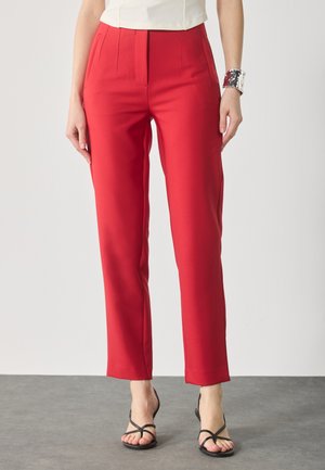 Red tailored trousers with a straight cut, featuring pleats at the front, and a smooth texture; paired with black strappy sandals.