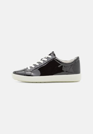 Zapatilla baja de charol negro con cordones blancos y suela gruesa de goma blanca.