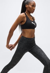 Γυναίκα με μαύρο αθλητικό τοπ και λεοπάρ κολάν τεντώνει τα χέρια πίσω από την πλάτη της, στέκεται με ανοιχτά πόδια σε απλό φόντο.