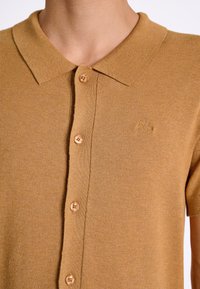 Lyhythihainen polo-paita vaaleanruskeasta neulekankaasta. Ominaisuuksiin kuuluu klassinen kaulus, viisi nappia ja brodeerattu logo rinnassa.