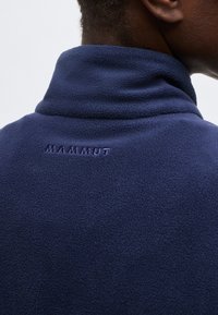 Κοντινό πλάνο ενός ατόμου που φοράει μπλε σκούρο μπουφάν από φλις με ψηλό γιακά και το λογότυπο "MAMMUT" κεντημένο στο πίσω μέρος.