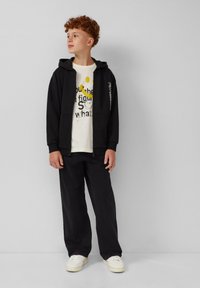 Adolescente con cabello rizado rojo, vistiendo una sudadera negra con cremallera, una camiseta gráfica blanca, pantalones negros y zapatillas blancas, de pie contra una pared sencilla.