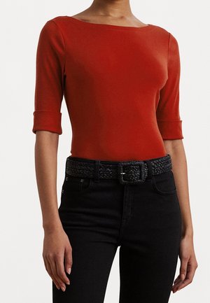 Vrouw draagt een aansluitend rood topje met boothals en driekwart mouwen en een zwarte broek met een geweven zwarte riem.