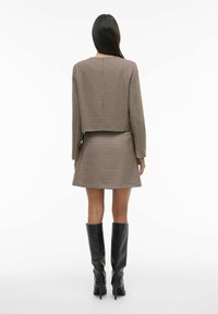 Skraćena jakna s uzorkom pepita u nijansama žućkasto-smeđe boje u kombinaciji s pripadajućom suknjom. Crne čizme do koljena upotpunjuju odjevnu kombinaciju.