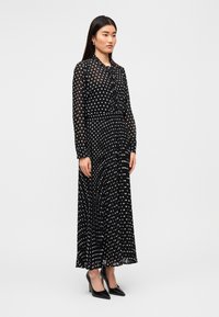 Μαύρο μακρύ φόρεμα με λευκές πουά, μακριά μανίκια, εφαρμοστή μέση και φουσκωτή φούστα. Συνδυάζεται με μαύρα παπούτσια με ψηλό τακούνι. Λεπτό, διαφανές ύφασμα.