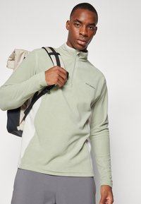Pull en polaire vert clair avec une fermeture éclair à mi-hauteur, présentant des panneaux latéraux blancs et un logo. Assorti à un sac à dos à carreaux beiges.