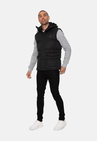 Schwarze Pufferweste mit Kapuze, die zwei Fronttaschen aufweist. Trägt eine graue Langarmbluse und kombiniert mit schwarzen Hosen und weißen Sneakers.