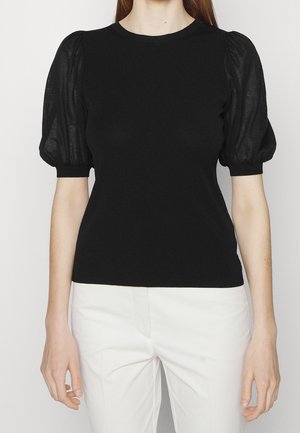 Vrouw draagt een zwart topje met korte mouwen en opgeblazen doorschijnende mouwen, en witte broek, staand tegen een effen achtergrond.