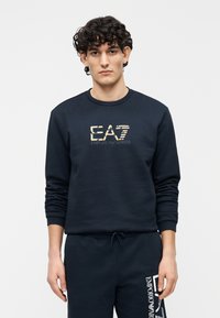 Tumesinine dressipluus ümarate kaeluse ja pikkade varrukatega, kuldse brändilogoga. Valmistatud pehmest kangast, millel on soonikkoes varrukasuu ja alumine äär.