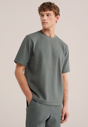 Mladý muž s kučeravými vlasmi oblečený v zladenom sivom tričku s krátkym rukávom a nohaviciach s textúrou, ruky v vreckách, pozerá sa bokom na jednoduchom pozadí.