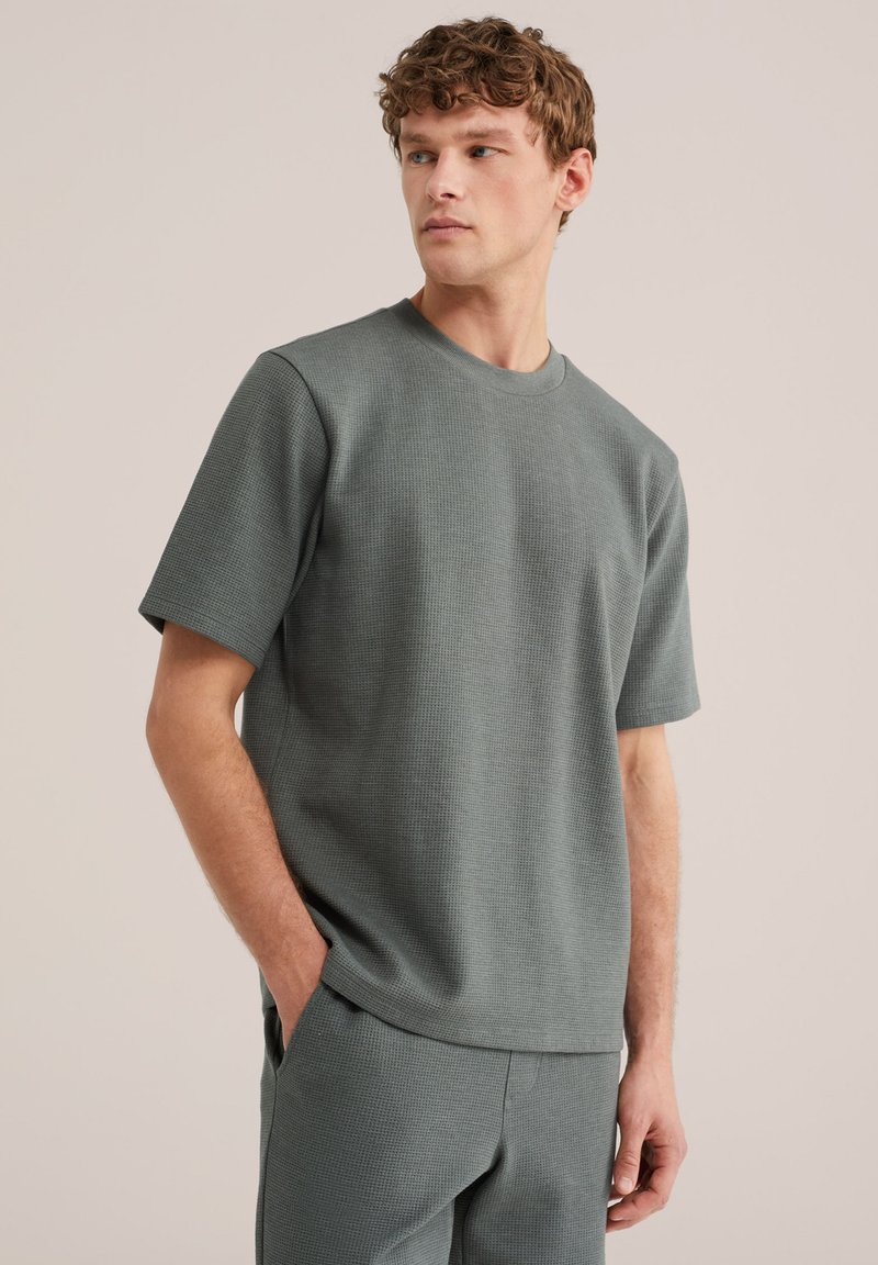 Fiatal férfi göndör hajjal, szürke, textúrált, rövid ujjú ingben és nadrágban, kezét zsebében tartva, oldalra néz egy egyszínű háttér előtt.